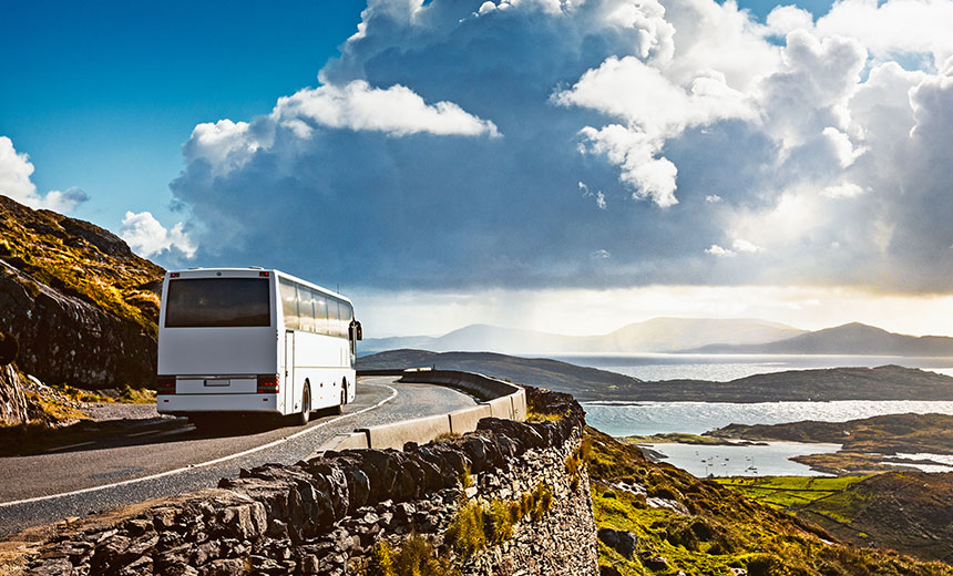 White coach driving along a winding coastal road with sea and mountains in the background