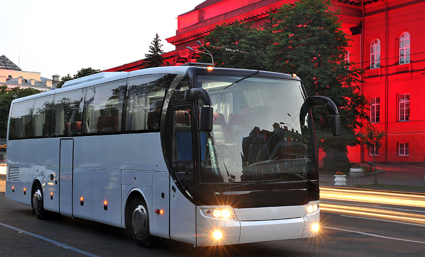 White coach travelling through a city street at dusk with headlights on and a red-lit building in the background.