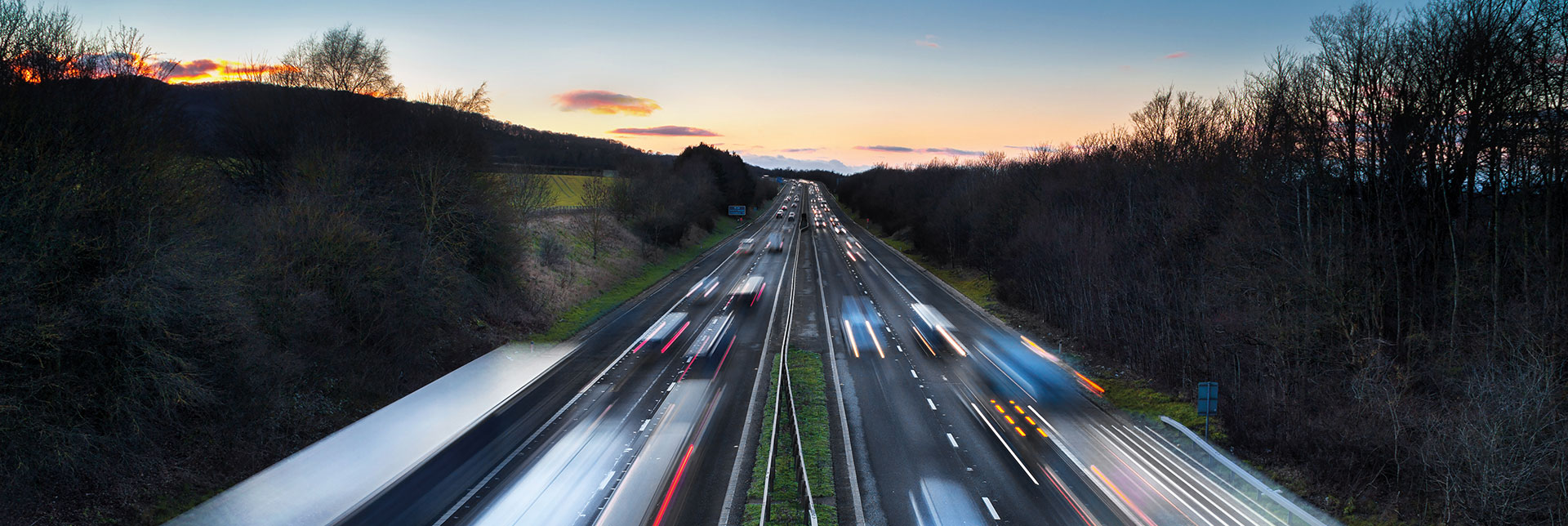 Motorway at dusk with trucks and cars travelling in both directions, headlights and taillights blurred by motion.