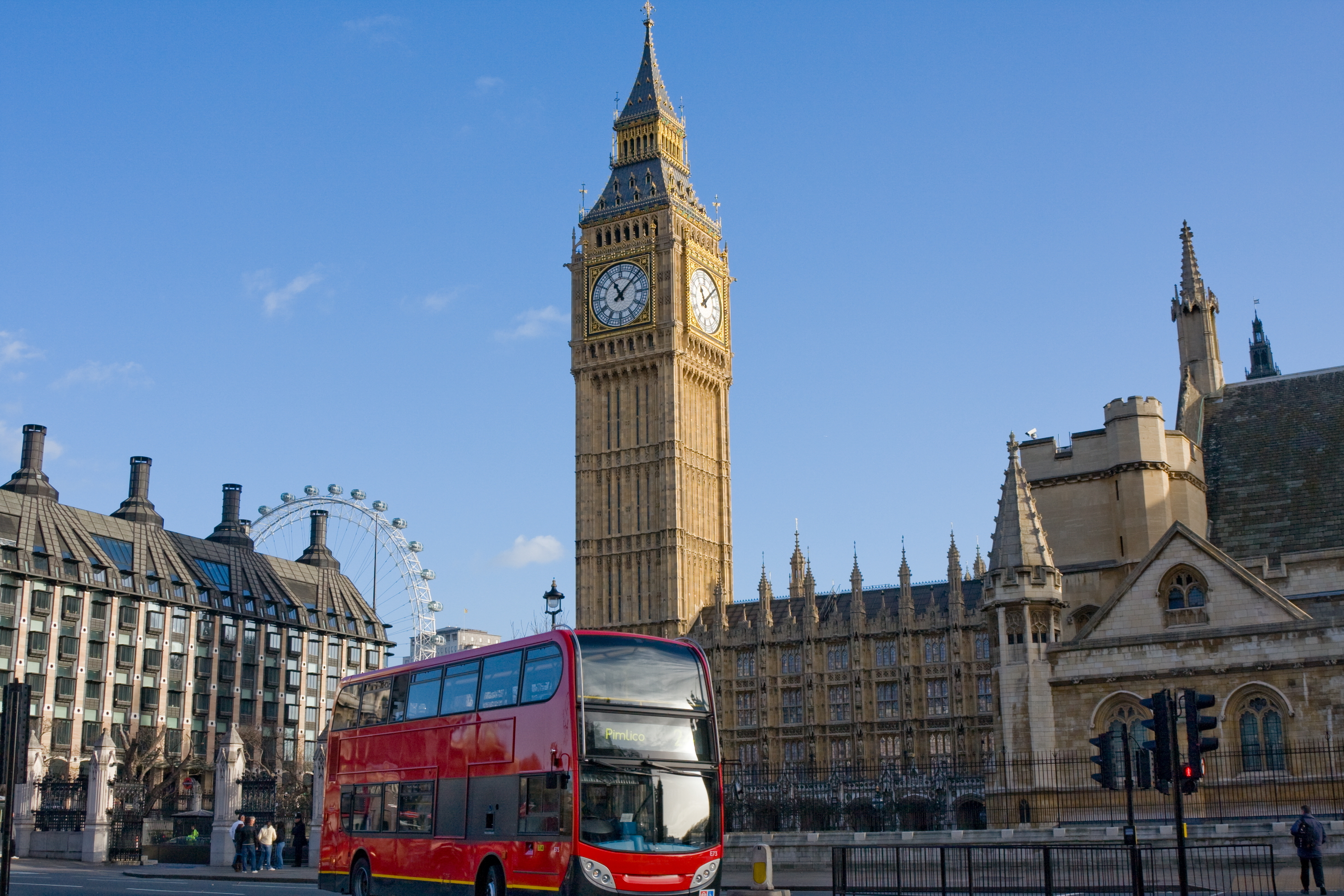 Red double-decker bus driving past the Houses of Parliament in London on a clear day.